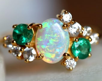 Könnte beinhalten: Ein goldener Ring mit einem ovalen Opal-Mittelstein, flankiert von zwei Smaragden und kleinen Diamanten. Der Opal zeigt schillernde Farben. Ein Schmuckstück.