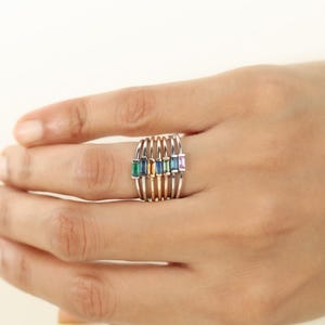 Op de afbeelding: Een veelkleurige edelsteenring met een uniek ontwerp. De ring heeft een reeks parallelle banden in zilver- en goudtinten, elk met een rechthoekige edelsteen in verschillende kleuren: groen, blauw, geel en roze. De ring wordt aan een hand gedragen.