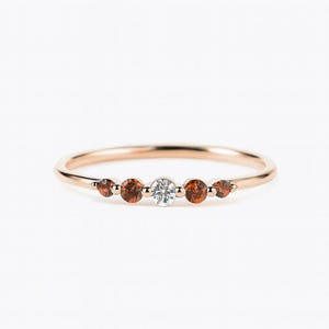May include: A rose gold ring featuring a central clear gemstone flanked by four smaller, round, reddish-brown gemstones. The ring has a delicate band and is set against a plain white background.