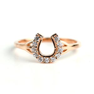 Könnte beinhalten: Roségoldener Hufeisenring mit klaren, rund geschliffenen Edelsteinen. Der Ring hat ein Hufeisen-Design, wobei die Edelsteine entlang der Kurve gesetzt sind. Das Band ist schlicht und dünn. Der Ring ist vor einem weißen Hintergrund platziert.