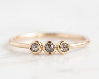 Puede incluir: Un anillo fino de oro con tres pequeñas piedras redondas facetadas de tono gris, engastadas individualmente en biseles. La banda es lisa y pulida, mostrando un diseño sencillo con las tres piedras dispuestas horizontalmente en el centro.