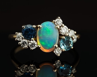 May include: A gold ring featuring an oval opal with blue and green iridescence, surrounded by a cluster of round diamonds, a dark blue sapphire, and a light blue gemstone. The ring is set against a black background.