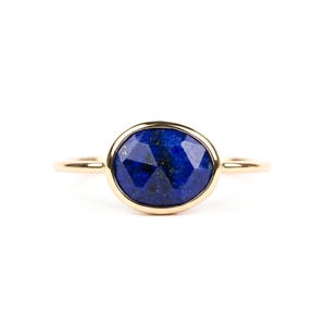 Puede incluir: Un anillo de oro con una piedra preciosa ovalada, facetada, de color azul oscuro. La piedra preciosa está engastada en un bisel de oro, y la banda del anillo es de oro fino y pulido. El anillo se muestra sobre un fondo blanco, resaltando el diseño de la joya.