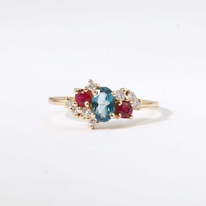 May include: A gold ring with an oval, light blue gemstone, two red gemstones, and small white gemstones. The ring has a delicate band and a cluster design. The gemstones are set in a cluster.