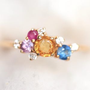 Puede incluir: Un anillo de oro con un grupo de gemas de colores. El anillo presenta una gema naranja grande y redonda en el centro, rodeada de piedras rosas, moradas, azules y blancas más pequeñas. El anillo tiene una delicada banda dorada.