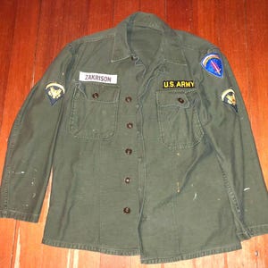May include: An olive green U.S. Army jacket with the name "ZAKRISON" on a name tag. The jacket has two chest pockets, patches on the sleeves, and the words "U.S. ARMY" above one pocket. The jacket is buttoned up and appears to be vintage.