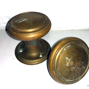 May include: Two round brass door knobs with a weathered, aged patina. One knob is attached to a circular mounting plate featuring two visible screw holes, while the second knob is detached and positioned to the right of the first piece.