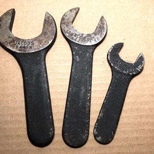 May include: Three vintage JACOBS wrenches of varying sizes, arranged on a brown surface. The wrenches are black with silver-colored heads, showing signs of wear. The largest wrench has "K440N" printed on it.