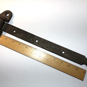 May include: A rusty, antique metal hinge with a decorative end and multiple screw holes. A wooden ruler, marked in inches, is placed alongside the hinge for scale. The hinge is likely for a door or cabinet.
