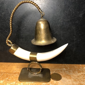 May include: A vintage brass bell with a curved white tusk-shaped support, accented with brass bands. The bell hangs from a twisted brass handle, and the tusk rests on a rectangular brass base. A small coin is next to the base.