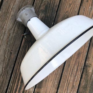 May include: A vintage white enamel light fixture with a black trim and a gray metal mounting. The fixture is angled, showing its curved shape. The background is weathered wooden planks.