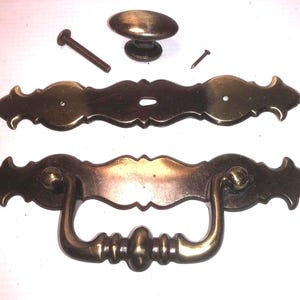 May include: A set of brass-toned metal furniture hardware including one oval knob, one decorative backplate with a central keyhole, one bail pull handle attached to a matching backplate, one threaded bolt, and one small metal nail.