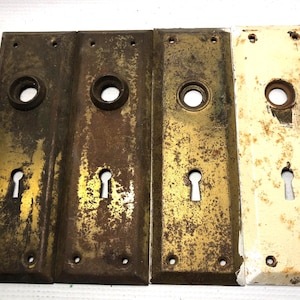 May include: Four antique door plates with keyhole openings. The plates are rectangular with a circular opening for the doorknob and three screw holes. The plates have a distressed, aged appearance with varying shades of gold, brown, and off-white.
