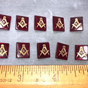May include: Ten square, burgundy-colored Masonic emblem pins. Each pin features a gold-colored symbol of a compass, square, and letter "G". The pins are arranged on a gray surface next to a wooden ruler.