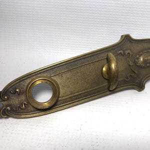May include: Antique brass door plate with a decorative design. The plate features a keyhole, a handle, and ornate detailing. The brass has a warm, aged patina, suggesting vintage craftsmanship. The overall shape is elongated with decorative edges.
