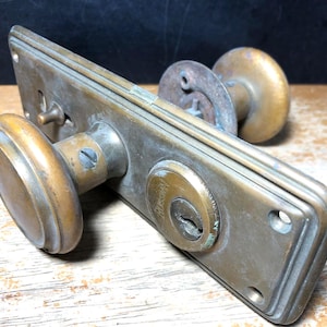 May include: Antique brass door hardware set, featuring a rectangular backplate, two round doorknobs, and a keyhole. The set shows signs of age and wear, with a patinaed finish. The hardware is designed for a door.