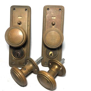 May include: Two sets of antique brass door hardware, each featuring a rectangular backplate with a round knob and a keyhole. Two additional matching round brass door knobs with spindles are positioned in the foreground, showing a worn metallic finish.