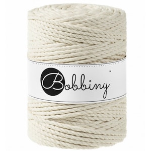 May include: A cylindrical spool of cream-coloured macramé cord. The cord is tightly wound and wrapped with a white label that reads "Bobbiny" in a cursive font. Suitable for various craft projects.