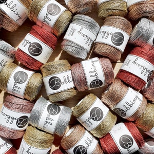May include: A collection of spools of colourful, metallic yarn with the brand name "Bobbiny" printed on each spool. The yarn is 3mm thick and is available in a variety of colours, including gold, silver, and red.