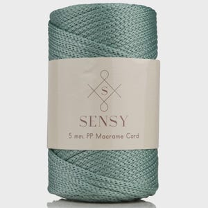 May include: A roll of sea green macrame cord. The label reads "SENSY" with the text "5 mm. PP Macrame Cord". The cord is tightly wound and has a textured appearance, suitable for various crafting projects.