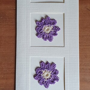 May include: A white card with three purple crocheted flowers with white centers. Each flower is framed in a white square.