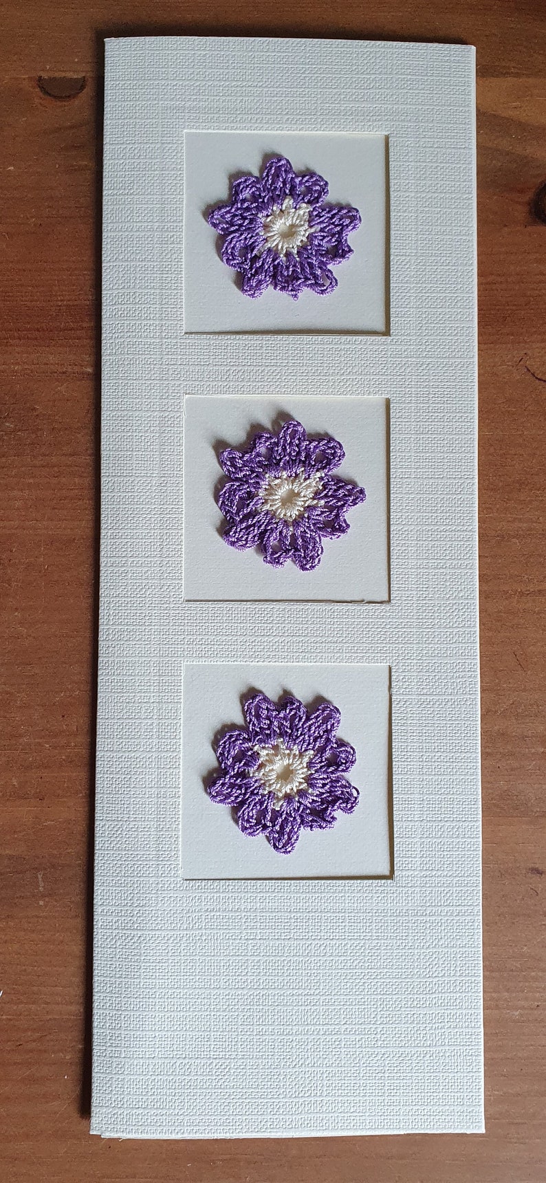 May include: A white card with three purple crocheted flowers with white centers. Each flower is framed in a white square.