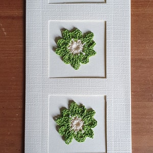 May include: A white card with three green crocheted flowers with white centers, each framed in a square.