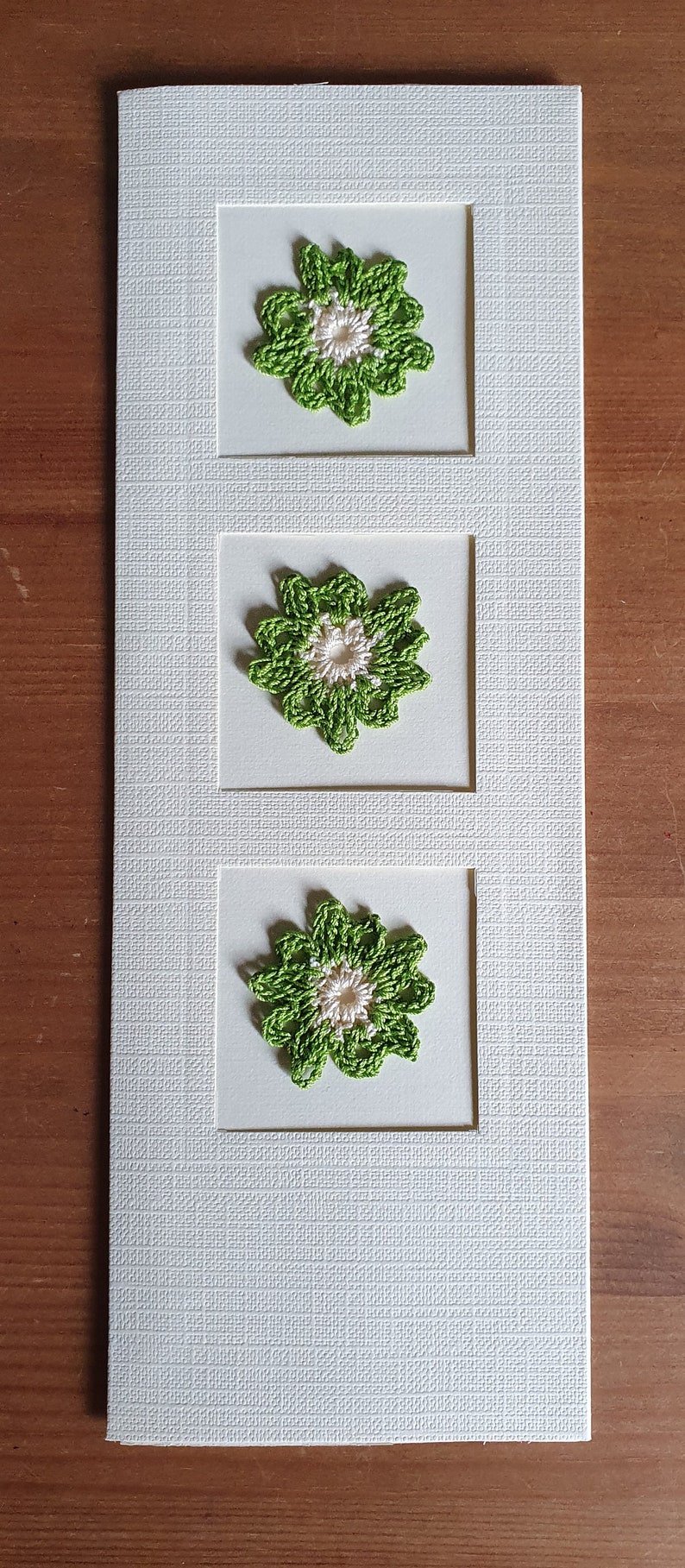 May include: A white card with three green crocheted flowers with white centers, each framed in a square.