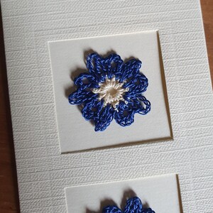 May include: A white card with two blue and white crocheted flowers. The flowers are framed in white.