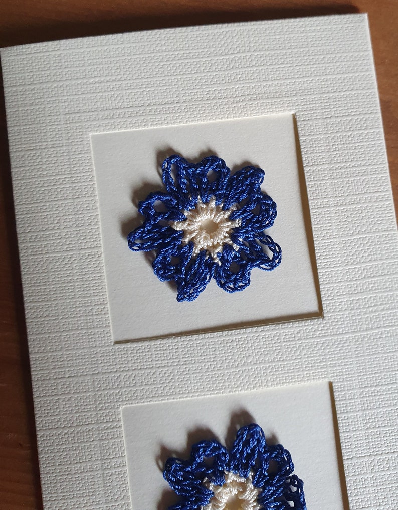 May include: A white card with two blue and white crocheted flowers. The flowers are framed in white.
