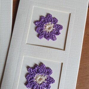 May include: A white card with two purple crocheted flowers with white centers. The flowers are framed in white squares.