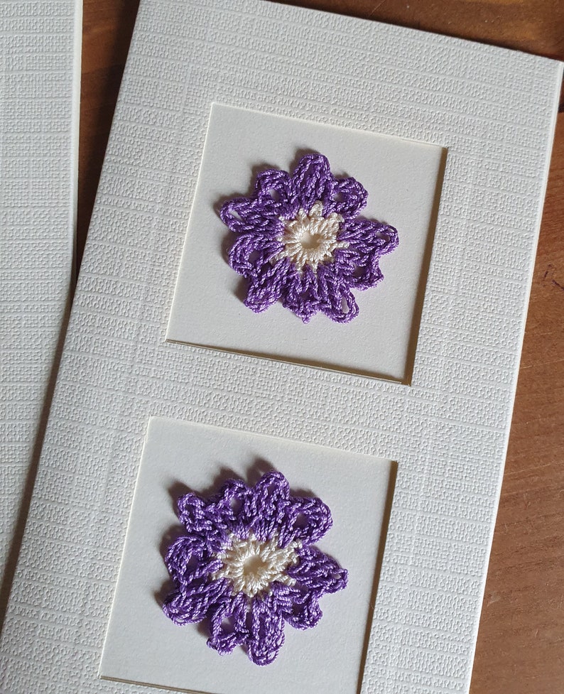 May include: A white card with two purple crocheted flowers with white centers. The flowers are framed in white squares.