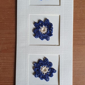 May include: A white card with three blue and white crocheted flowers in frames.