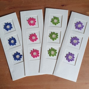 May include: Four white cards with different colored crocheted flowers in each square. The flowers are pink, green, purple, and blue.