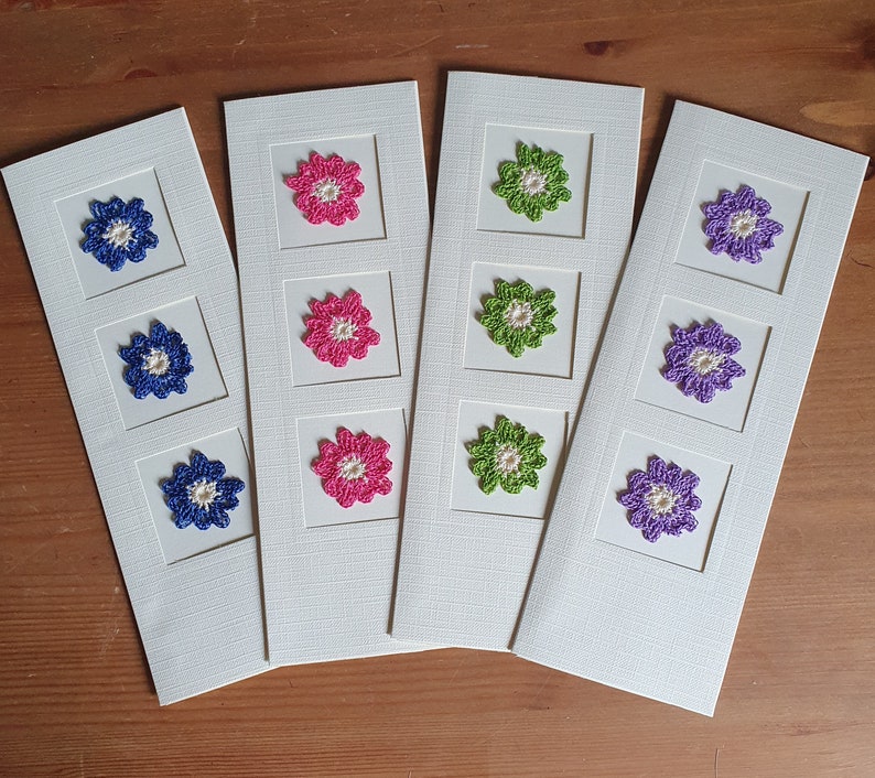 May include: Four white cards with different colored crocheted flowers in each square. The flowers are pink, green, purple, and blue.