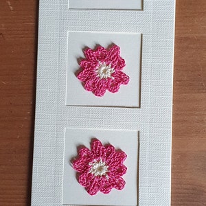 May include: A white card with three pink crocheted flowers in square frames. The flowers have a white center.