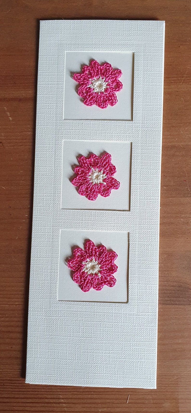 May include: A white card with three pink crocheted flowers in square frames. The flowers have a white center.