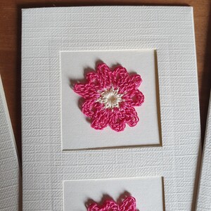 May include: A white card with two pink crocheted flowers. The flowers have a white center and are framed in white.