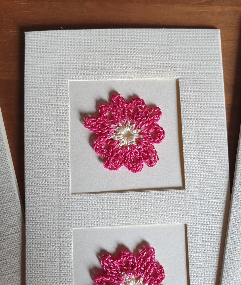 May include: A white card with two pink crocheted flowers. The flowers have a white center and are framed in white.