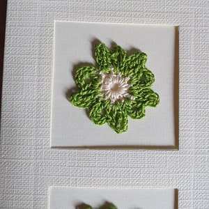 May include: A white card with two green and white crocheted flowers. The flowers are framed in white.