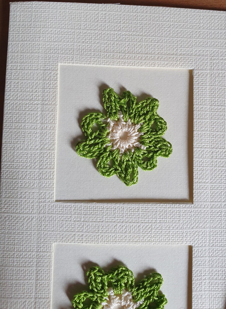 May include: A white card with two green and white crocheted flowers. The flowers are framed in white.