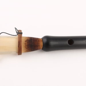 Könnte beinhalten: Eine schwarze Holzduduk mit einem braunen Holzmundstück und einem weißen Rohr. Die Duduk hat vier Fingerlöcher und eine kleine Schnur, die am Mundstück befestigt ist.