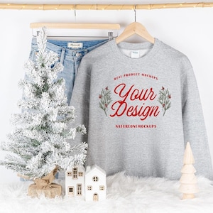 May include: A gray sweatshirt with the text "REAL PRODUCT MOCKUPS Your Design NATUREONEMOCKUPS" printed on the front. The sweatshirt is hanging on a wooden hanger and is surrounded by a white fluffy surface, a small snowy Christmas tree, and two small white houses.