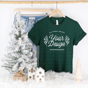 May include: A dark green t-shirt with a white design that says "Your Design" and "Real Product Mockups" and "NatureOneMockups". The t-shirt is hanging on a wooden hanger and is displayed on a white fluffy surface with a small white house and a small pine tree.