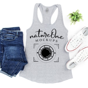 Op de afbeelding: Een grijs tanktop met de tekst "natureone MOCKUPS" en een afbeelding van een camera lens in een vierkant frame. Het tanktop ligt op een wit oppervlak met een paar witte sneakers en een kleine plant in een pot.
