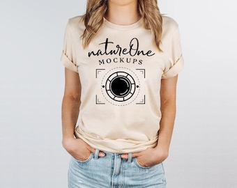 May include: A beige short-sleeved t-shirt featuring a black graphic print on the chest. The text reads natureOne MOCKUPS above a circular camera lens icon framed by dashed lines and corner brackets. The sleeves are rolled up at the edges.
