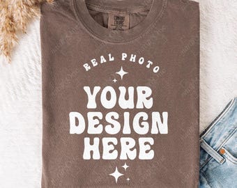Könnte beinhalten: Ein braunes kurzärmeliges T-Shirt mit Rundhalsausschnitt. Auf der Vorderseite befindet sich der weiße Schriftzug REAL PHOTO YOUR DESIGN HERE in geschwungener und gestapelter Anordnung, ergänzt durch kleine vierzackige Sternmotive um den Text.