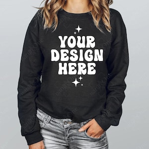 Puede incluir: Una sudadera negra de manga larga con el texto blanco YOUR DESIGN HERE en una fuente ondulada y negrita, centrada en el pecho. El texto está flanqueado por pequeños iconos de estrellas de cuatro puntas arriba y abajo, con pantalones vaqueros claros.