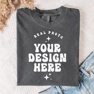 May include: A charcoal grey crew neck t-shirt featuring the white text "REAL PHOTO YOUR DESIGN HERE" in a bold, wavy font. The design is accented with small four-pointed star icons above and below the text, centred on the chest area.