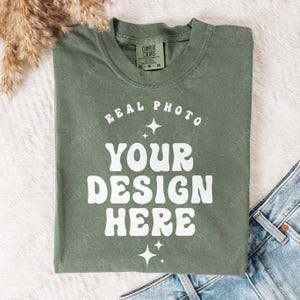 May include: An olive green short-sleeve t-shirt with a crew neckline. The front features white text that reads REAL PHOTO YOUR DESIGN HERE, arranged in a curved and stacked layout, accented by small four-pointed star icons above and below the text.